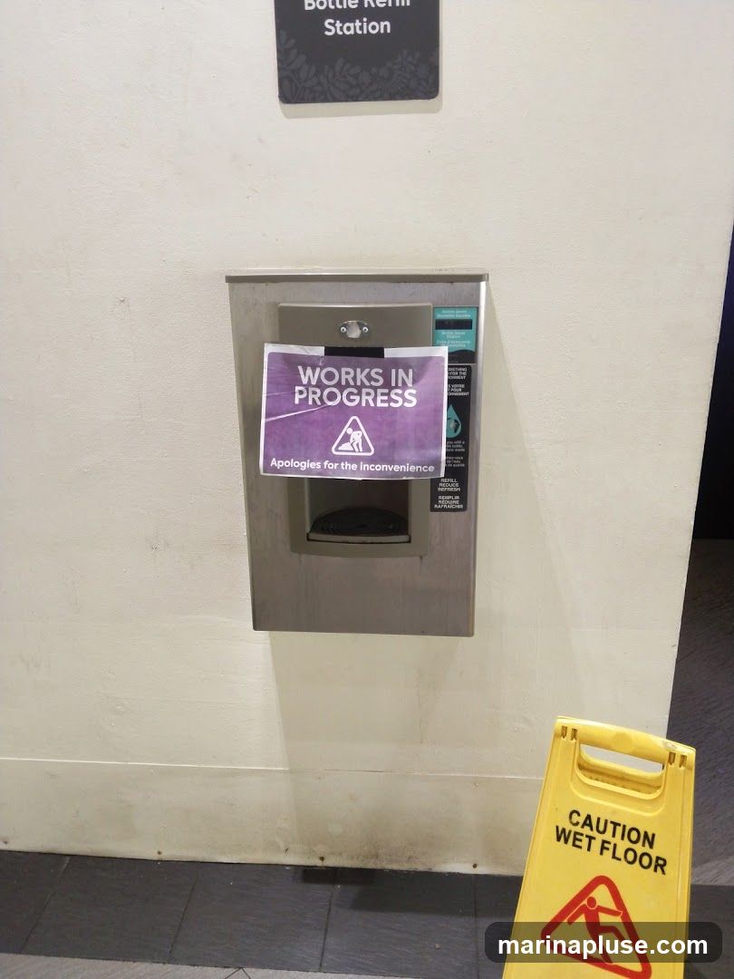 A bottle refill station is covered with a "WORKS IN PROGRESS" sign. Below it, a yellow "CAUTION WET FLOOR" sign is visible.