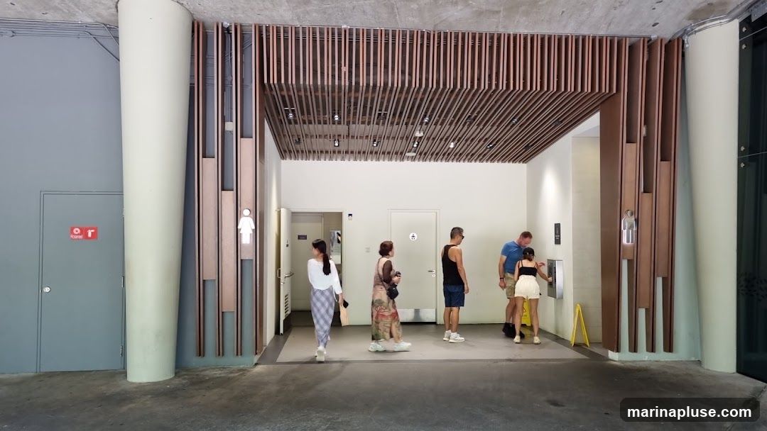 People are seen near the entrance of public restrooms. There are signs indicating men's and women's restrooms. The area has a modern design with wooden slats on the ceiling and walls.