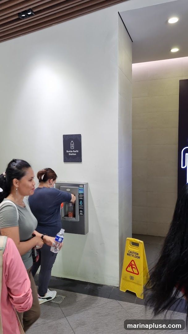 A bottle refill station is mounted on a white wall. A woman is using the station to refill a bottle. A yellow "Caution Wet Floor" sign is visible to the right.