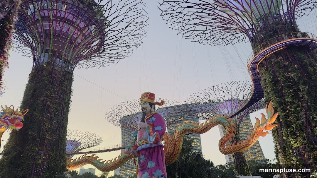 A festive display featuring large, illuminated Supertrees adorned with greenery and lights. In the foreground, a statue of a traditional Chinese deity stands prominently, flanked by a long, golden dragon sculpture. In the background, the iconic Marina Bay Sands hotel is visible. The sky is a soft gradient of orange and blue, suggesting dusk or dawn.