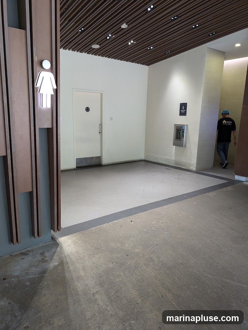 A view of a restroom entrance with a female symbol sign on the left. There is a white door with a circular symbol and a silver handle. To the right, there is a wall-mounted dispenser and a sign that reads "Emergency Station". A person wearing a black t-shirt and blue hairnet is walking away from the camera. The floor is tiled and concrete.