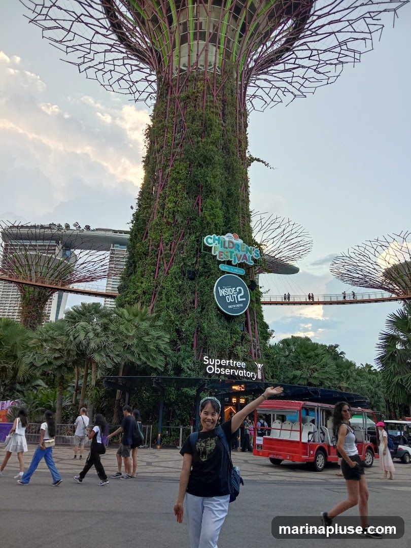A woman poses for a photo in front of the Supertree Observatory at Gardens by the Bay. Other visitors are seen walking around the area, and a red tourist vehicle is parked nearby. The Singapore skyline is visible in the background.