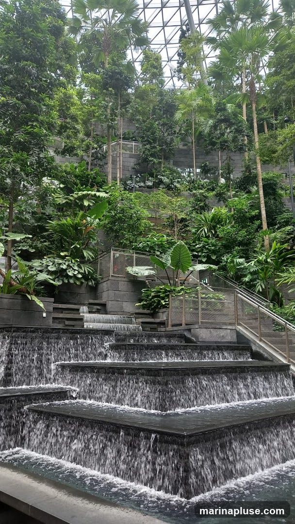 A multi-tiered indoor waterfall cascades down dark stone steps surrounded by lush greenery and tall palm trees. The scene is enclosed by a geodesic dome structure, suggesting an indoor botanical garden or atrium. Metal railings and stairs are visible on the right side, indicating pathways for visitors.