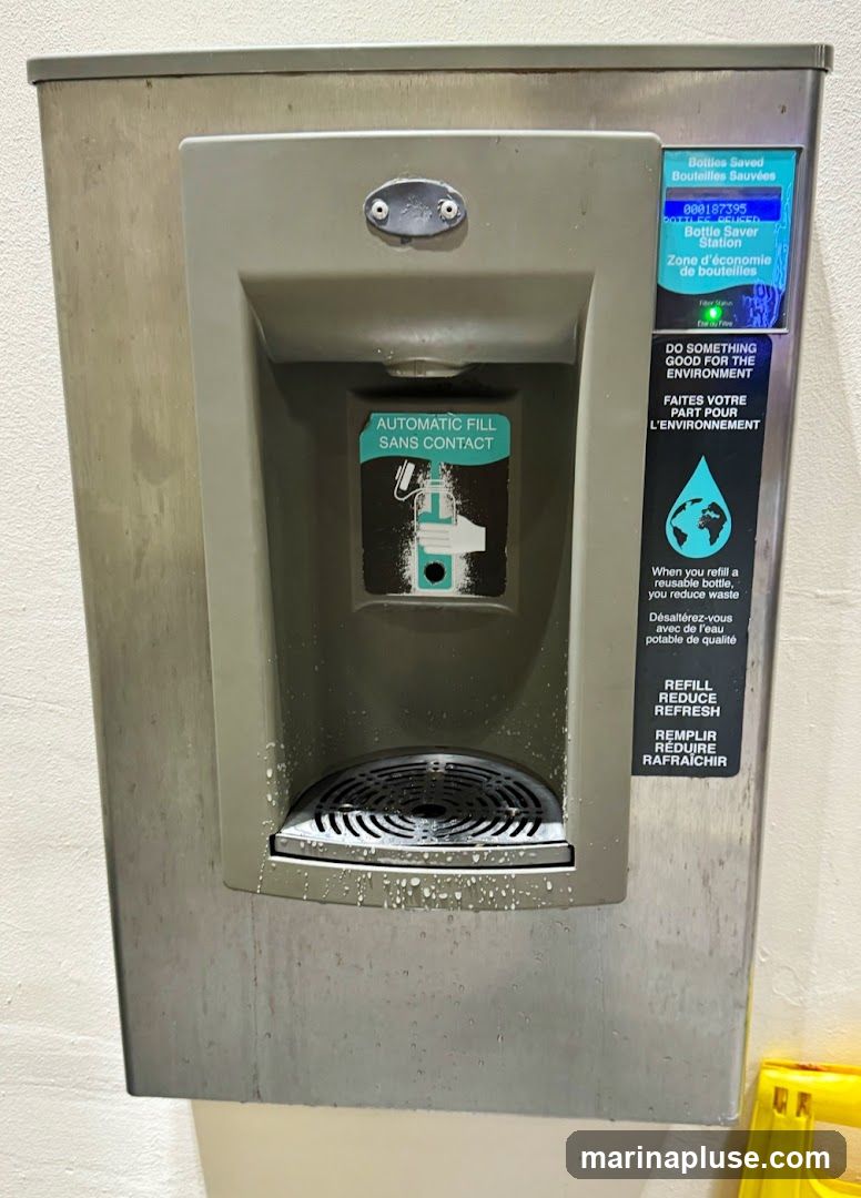 A stainless steel bottle refill station with a digital display showing "Bottles Saved" and a count of 000187395. It features an "Automatic Fill Sans Contact" instruction with an illustration. A sticker encourages environmental action with the text "Do Something Good for the Environment" and "When you refill a reusable bottle, you reduce waste." Another sticker reads "Refill Reduce Refresh." The station appears to be in a public area, possibly a school or transit hub, with some water splashes visible around the dispensing area.