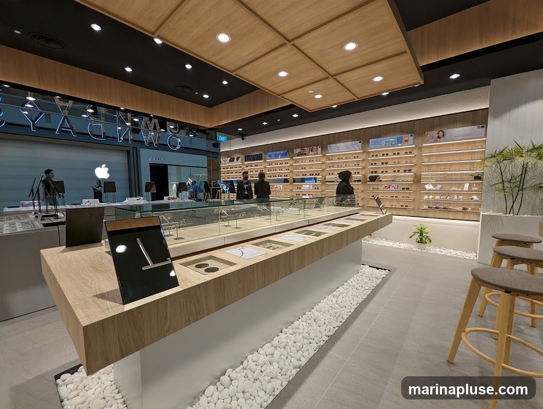 The interior of a modern eyewear store with a clean and minimalist design. The store features a long display counter with glass cases showcasing eyeglasses. Behind the counter, shelves are filled with various eyewear frames, illuminated by soft lighting. In the background, there are signs for other businesses, including Apple and Zara. The overall ambiance is bright and organized, with wooden accents and white pebble landscaping adding a touch of warmth and nature.