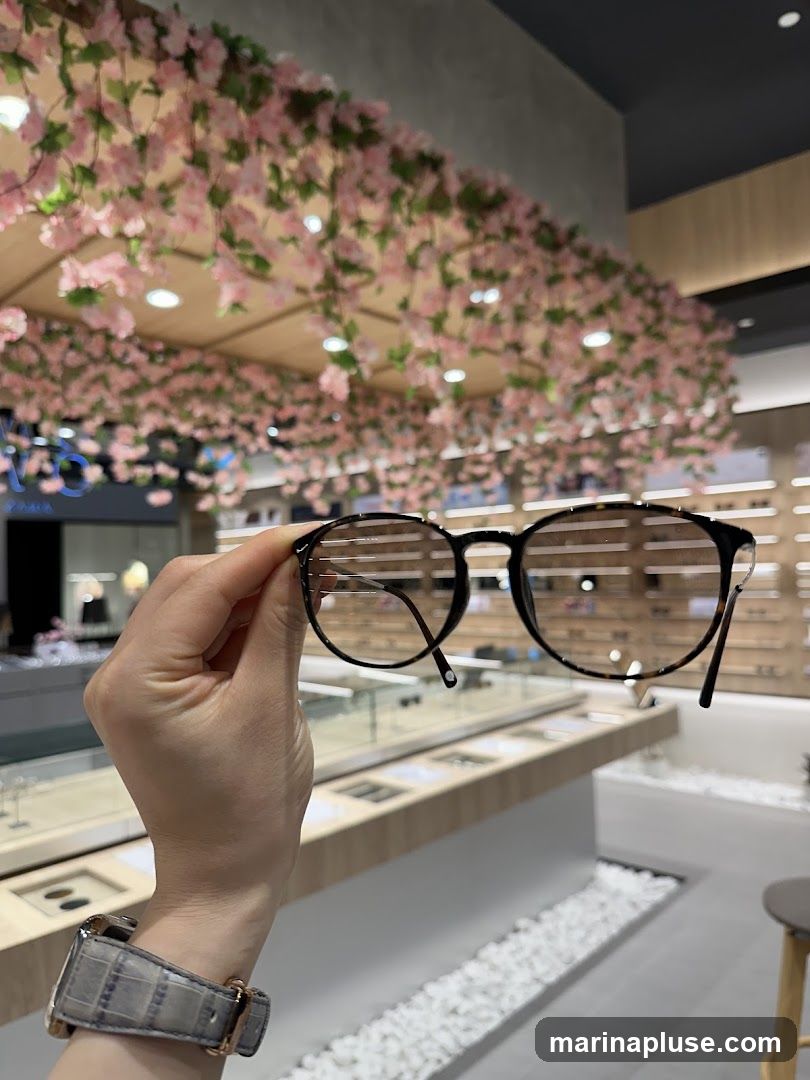 A hand holds up a pair of eyeglasses in front of a display of more glasses in a store. The store has a modern design with wooden accents and decorative pink flowers hanging from the ceiling.