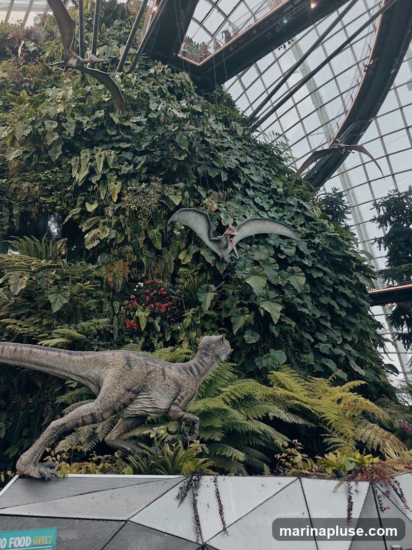 A realistic model of a velociraptor dinosaur is positioned in the foreground, with its body angled to the right. Behind it, a lush, vertical garden is densely packed with various green plants and ferns. Two pterodactyl models are suspended in the air, one in the mid-ground and another further back, appearing to fly amongst the foliage. The scene is enclosed by a large, geodesic glass dome structure, with visible metal beams and a walkway overhead where people can be seen. A sign with the text "NO FOOD ONLY" is partially visible at the bottom left.