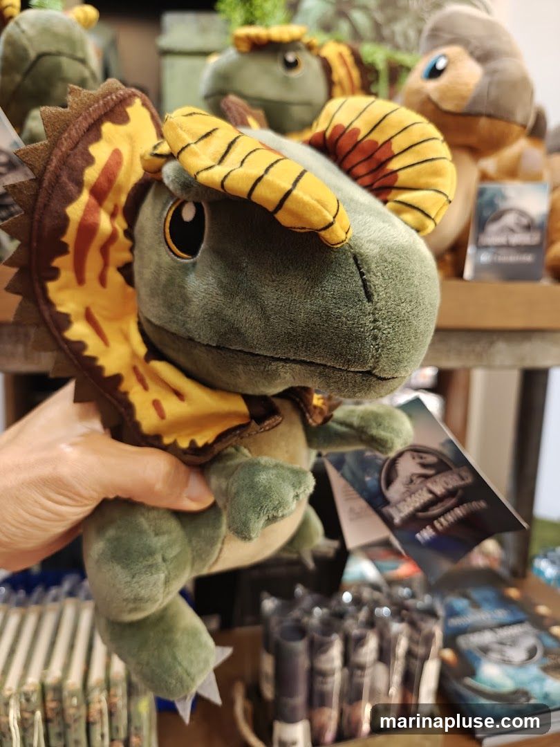 A hand holds a plush toy of a dinosaur with a frill around its neck. In the background, more dinosaur plush toys and merchandise are visible, including items with the Jurassic World logo.