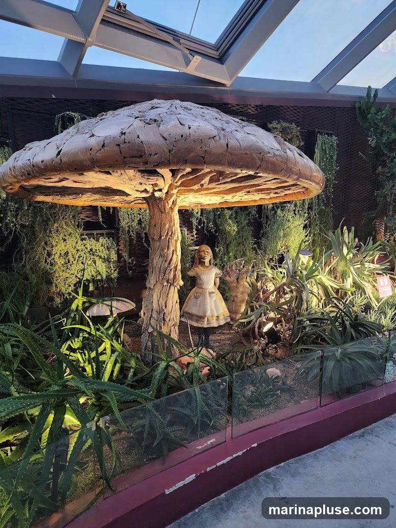 A whimsical display featuring a large, textured mushroom sculpture with a tree-like trunk. A figurine of Alice in Wonderland stands beneath the mushroom cap. The scene is surrounded by lush green plants, including aloe vera, and is illuminated by spotlights. The display is enclosed by a glass barrier.