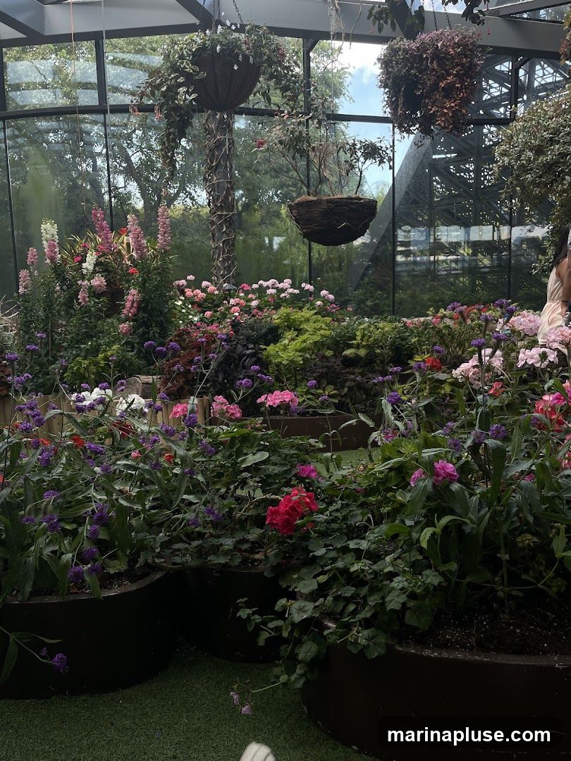 A lush indoor garden filled with a variety of colorful flowers and plants. Hanging baskets are suspended from the ceiling, and large planters hold a dense arrangement of blooms. The space is enclosed by glass walls, offering views of trees and architectural structures outside.
