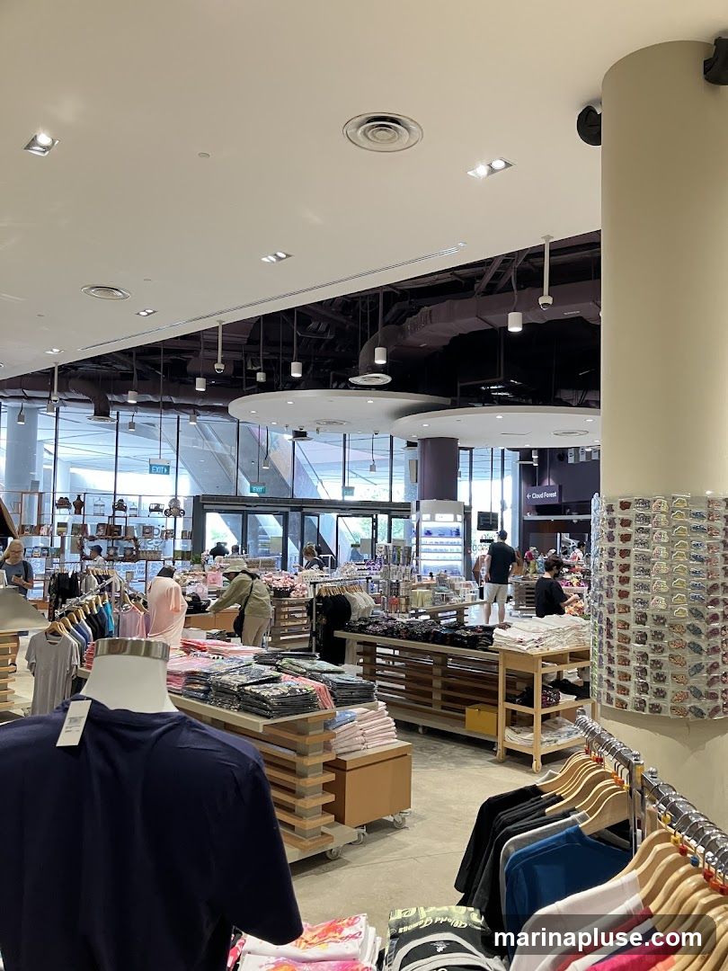 A view inside a brightly lit retail store, showcasing racks of t-shirts, folded apparel, and various merchandise displayed on shelves and tables. Mannequins are visible, dressed in sample clothing. In the background, customers browse and an exit sign is illuminated. The store appears to be a gift shop or souvenir store, possibly within a larger attraction, given the signage for "Cloud Forest" visible in the distance.