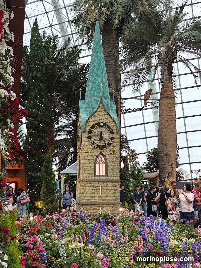 A decorative clock tower with a green steeple stands amidst a vibrant display of colorful flowers. Palm trees and other greenery surround the tower, and a glass dome structure is visible in the background. Several people are observing the display and taking pictures. A decorative bird figure is perched on a branch near one of the palm trees.
