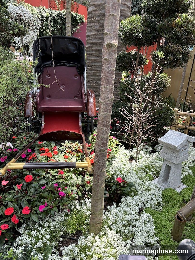 A maroon rickshaw with a black canopy sits amidst a lush garden. The garden features a variety of plants, including red and pink impatiens, white alyssum, and green ground cover. A small stone lantern and bamboo accents add to the oriental theme. Palm trees and other foliage create a dense backdrop.