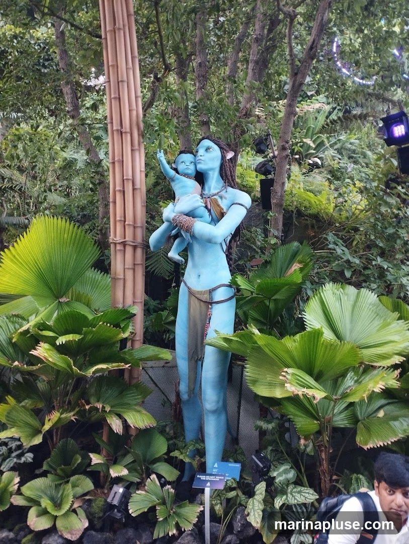 A statue of a blue-skinned Na'vi woman holding a baby, set in a lush, green jungle environment with bamboo stalks and large tropical leaves. A sign at the base of the statue reads "PLEASE DO NOT TOUCH THE STATUE.". A man is visible in the lower right corner of the image.
