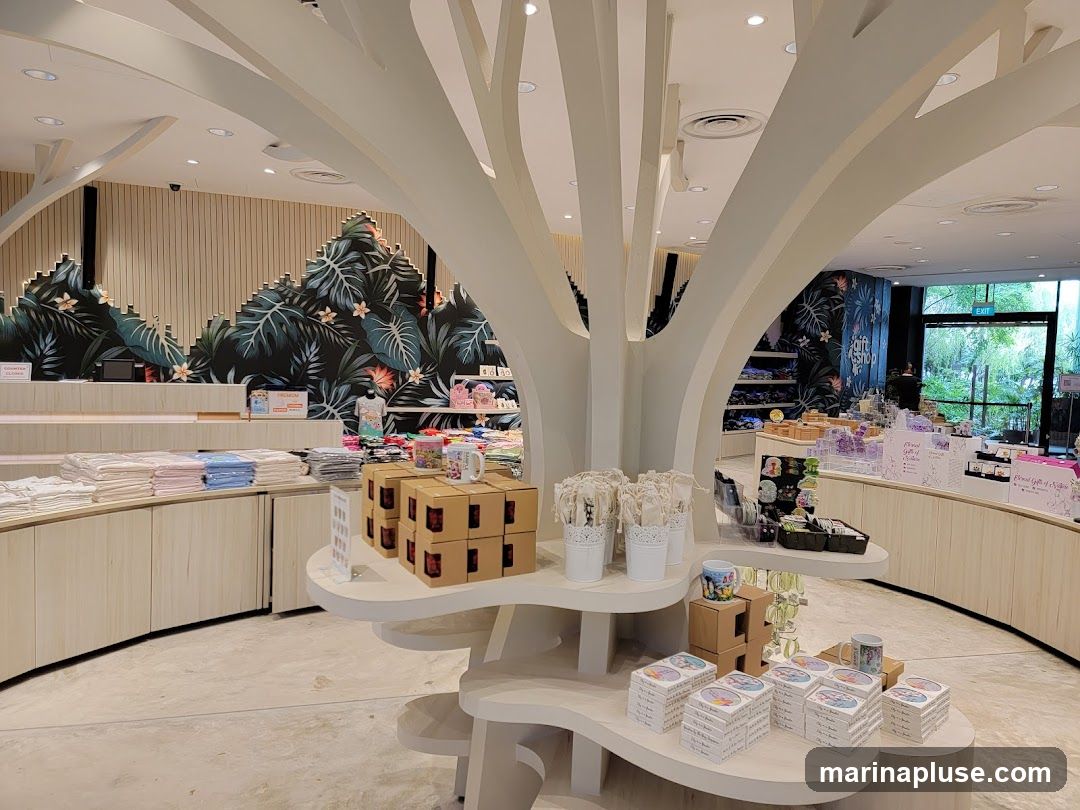 A gift shop interior with displays of merchandise. In the foreground, there are stacks of boxes and mugs on a white tree-like display. To the left, folded textiles are arranged on shelves. The background features tropical-themed wallpaper and more product displays. An exit sign is visible through a glass door in the distance.