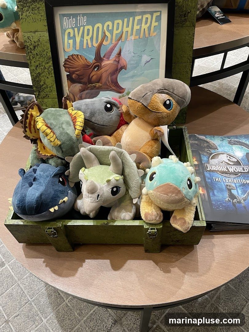 A collection of plush dinosaur toys are displayed in a green wooden crate. Behind the crate, a framed poster reads "Ride the GYROSPHERE" and features an illustration of a Triceratops. To the right of the crate, a brochure or book with the title "JURASSIC WORLD THE EXHIBITION" is visible.