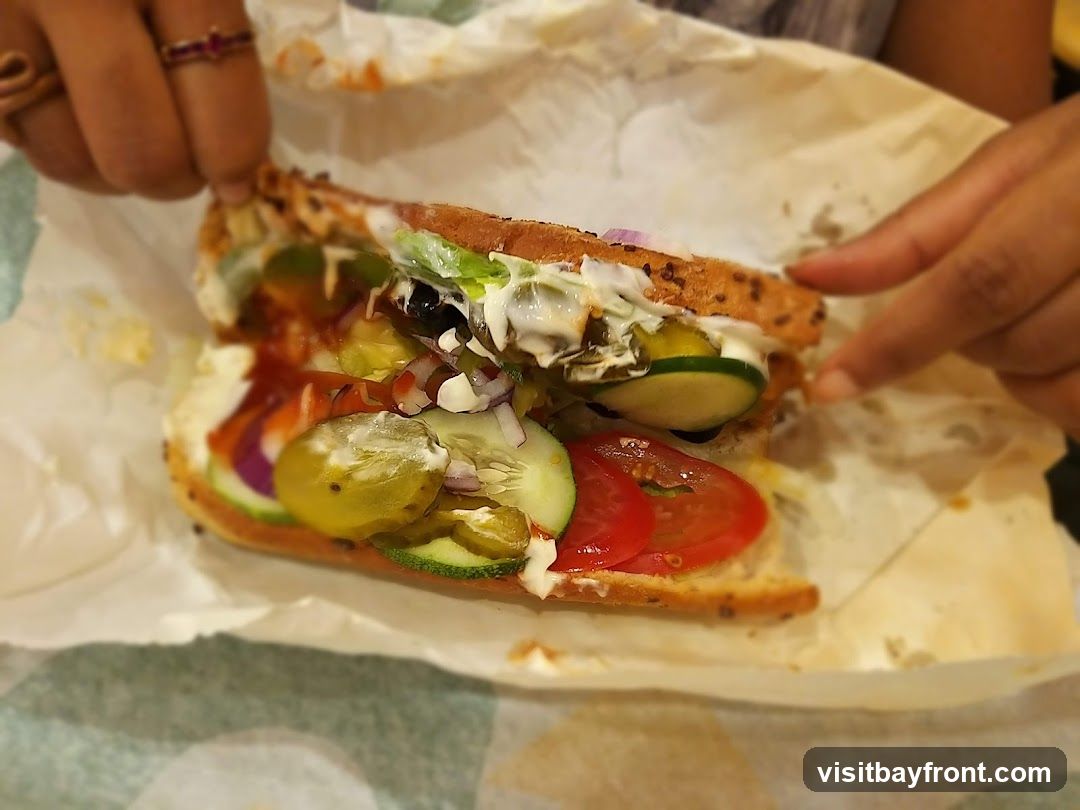 A close-up shot of a sub sandwich filled with various vegetables and sauces, wrapped in paper. The sandwich has lettuce, tomatoes, cucumbers, onions, and pickles. Two hands are visible, one holding the sandwich from the left and the other adjusting it from the right. The background is blurred, showing a patterned surface.