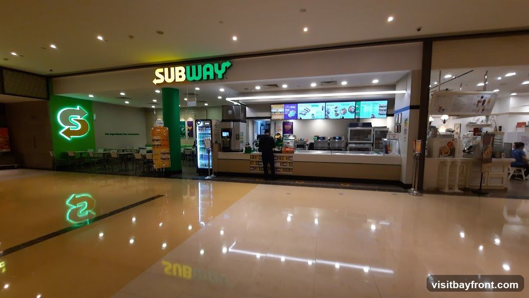 The exterior of a Subway restaurant located in a mall. The prominent Subway logo is visible above the counter, and a large green illuminated 'S' logo is on the left wall. Seating areas are visible inside the store. A person is standing at the counter, and another person is seated in the background. Digital menu boards display food items and prices.