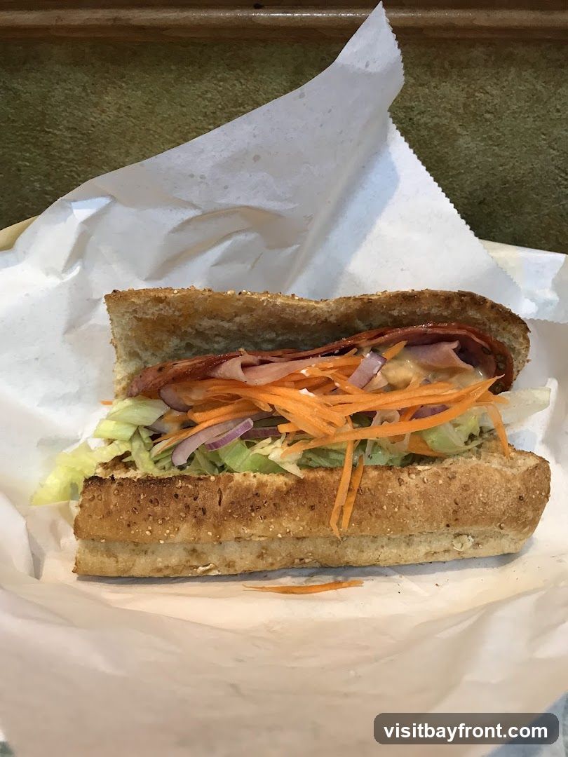A close-up shot of a sandwich on a whole wheat bun, filled with shredded carrots, lettuce, red onion, and meat, served in white paper.