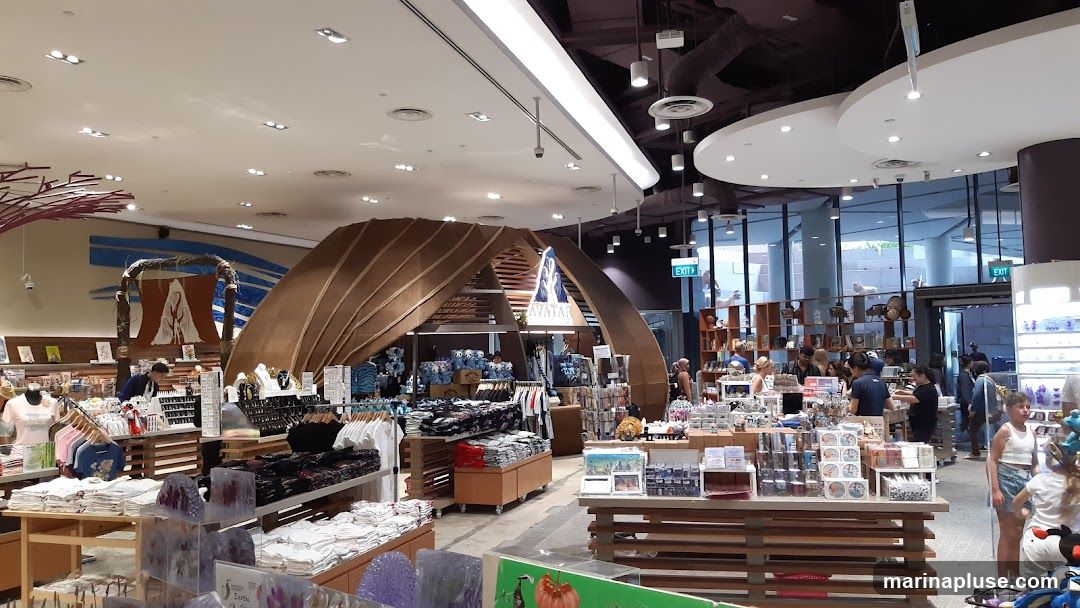 A wide shot of the interior of a gift shop, likely themed around the movie Avatar. The store is filled with merchandise displayed on shelves and racks. In the center, a prominent structure with the word "AVATAR" is visible. People are browsing the various items, including clothing, plush toys, and souvenirs. The lighting is bright, and the overall atmosphere is that of a busy retail environment.