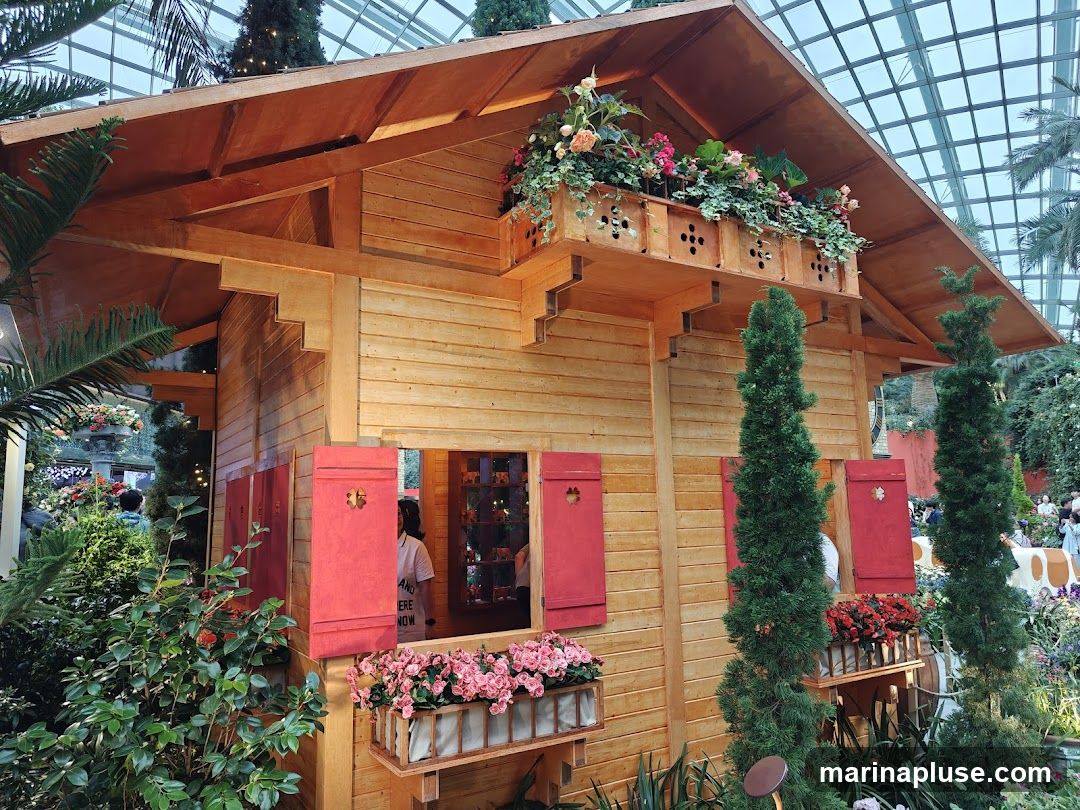 A charming wooden cabin-like structure is adorned with vibrant flowers in window boxes and on a balcony. Red shutters add a pop of color to the wooden facade. The structure appears to be part of a larger display, possibly in a botanical garden or exhibition, with lush greenery and other floral arrangements visible in the background. The overall scene evokes a rustic and picturesque ambiance.
