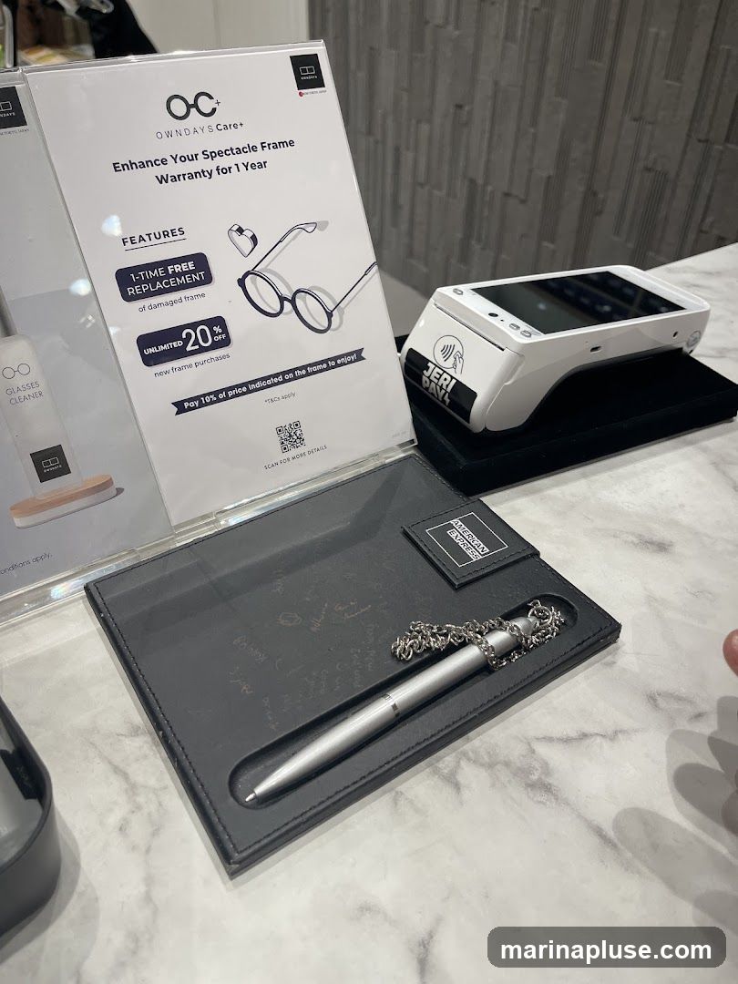 A promotional display for OWNDAYS Care+ offering a 1-year spectacle frame warranty. Features include a 1-time free replacement for damaged frames and unlimited 20% off new frame purchases. A payment terminal and a pen on a leather tray are also visible.