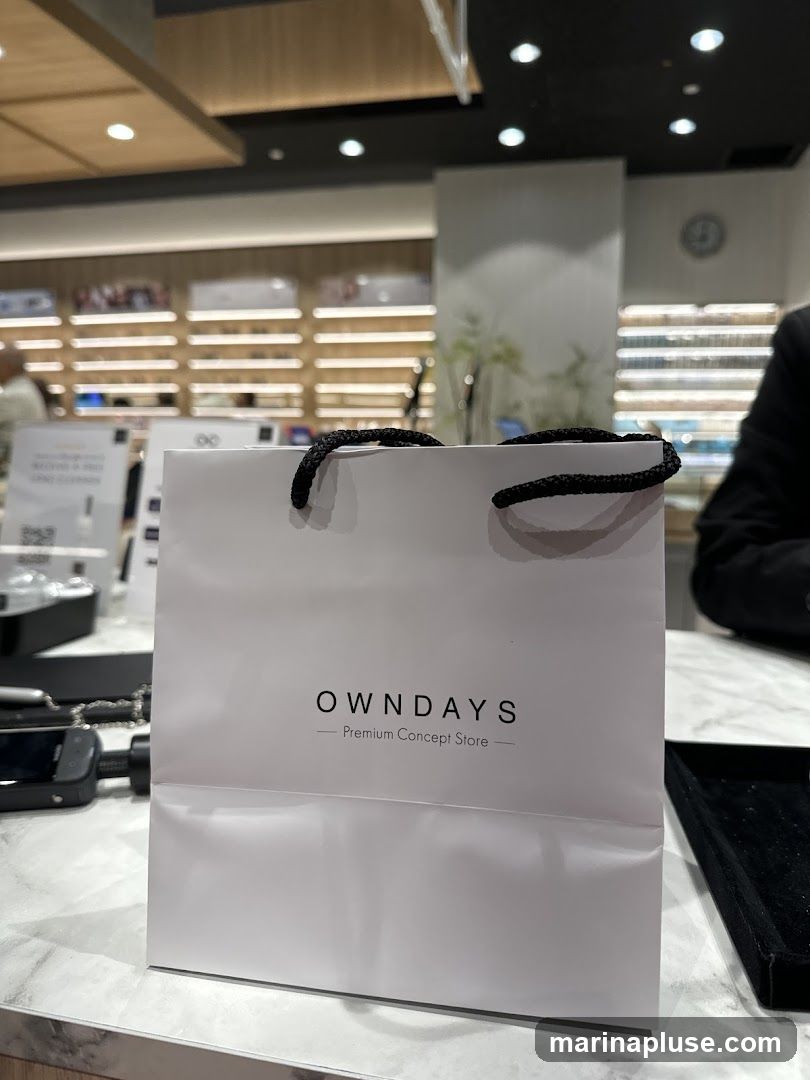A white shopping bag with black handles sits on a marble countertop. The bag has the OWNDAYS logo and the text "Premium Concept Store" printed on it. In the background, shelves filled with eyeglasses are visible, suggesting the bag is from an optical store.