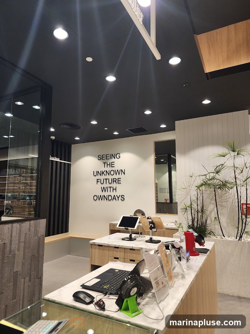 The image shows the interior of a modern retail space. There are several display counters with tablets and laptops, suggesting an electronics or technology store. A prominent wall features the text "SEEING THE UNKNOWN FUTURE WITH OWNDAYS" in large black letters. The space is well-lit with recessed ceiling lights and decorated with potted plants.