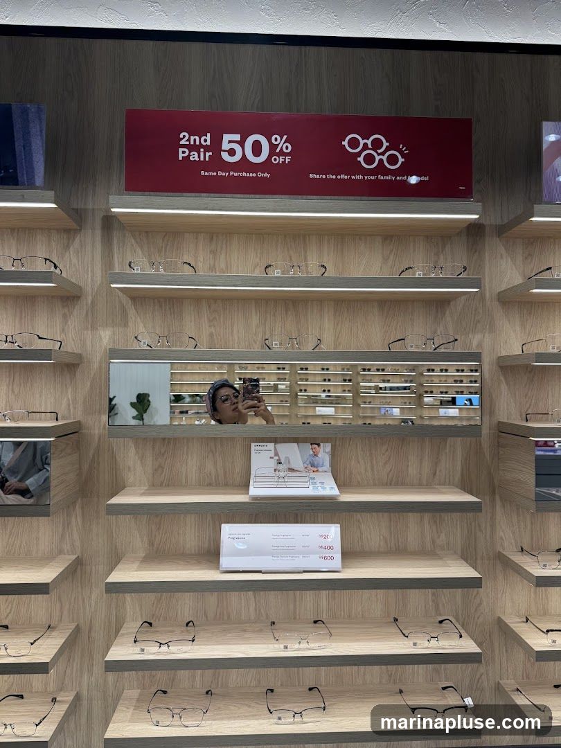 A wall display of eyeglasses on shelves. A red promotional banner at the top advertises "2nd Pair 50% OFF" with "Same Day Purchase Only". Below the banner, a mirror reflects a person taking a selfie. Further down, a price list shows options for progressive lenses with prices ranging from $200 to $600.