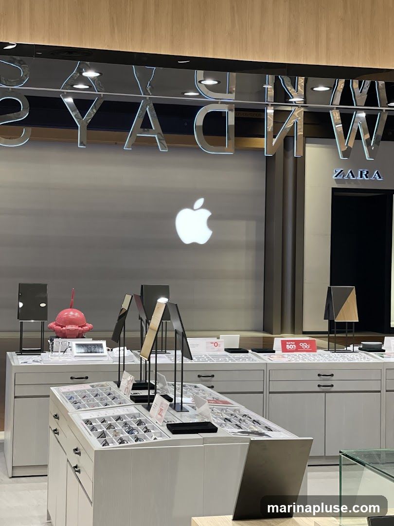 An interior view of a retail store, likely an Apple Store, with displays of eyeglasses and accessories. Prominent Apple logos are visible on the wall. Signage for Zara is also present. The display cases are filled with various eyewear options. Promotional signs indicating discounts are placed on the counters.