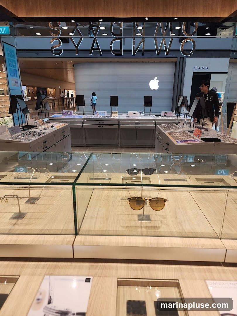 A well-lit interior of an eyewear store with displays of sunglasses and eyeglasses. In the background, there is an Apple store and a Zara store. The store name, reflected in the ceiling, appears to be OWNDAYS.