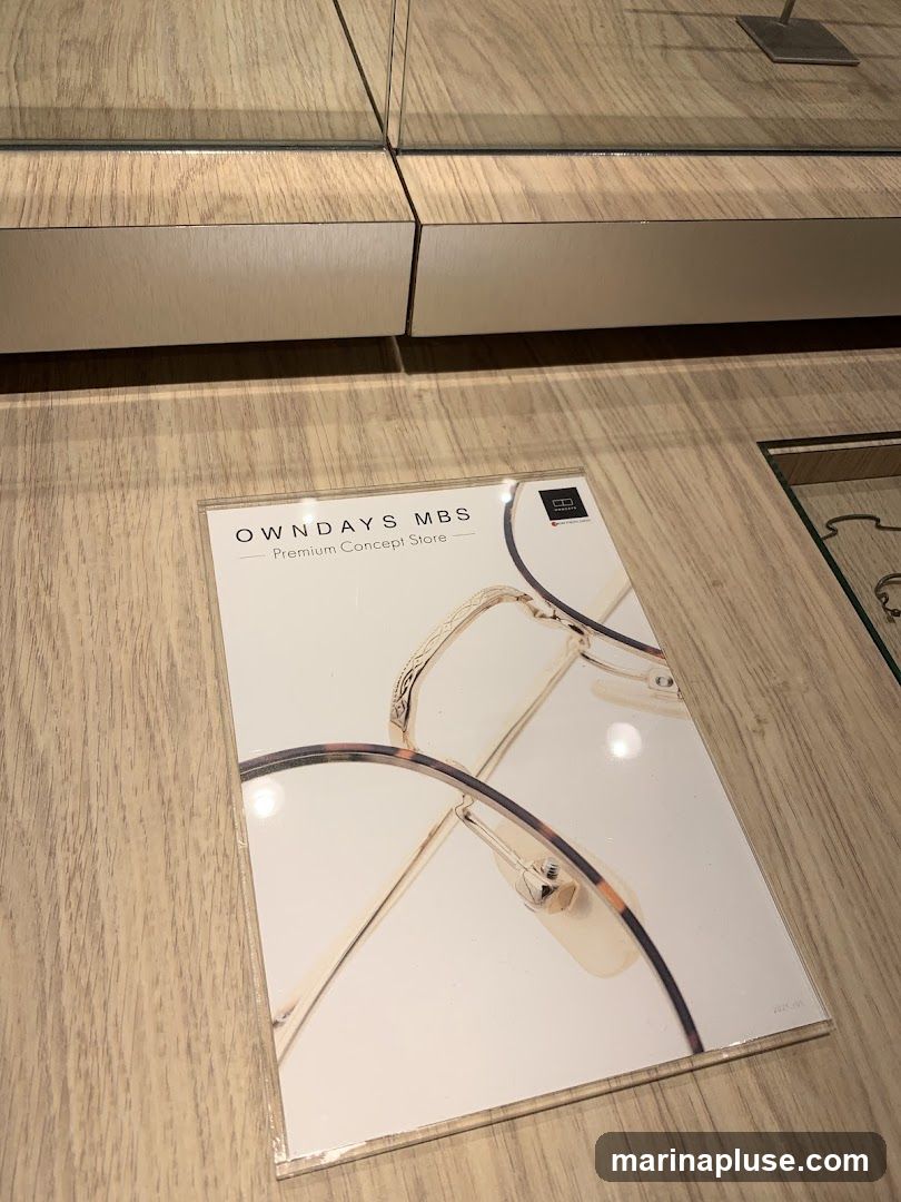 A brochure for OWNDAYS MBS Premium Concept Store is displayed on a wooden surface. The brochure features images of eyeglasses.