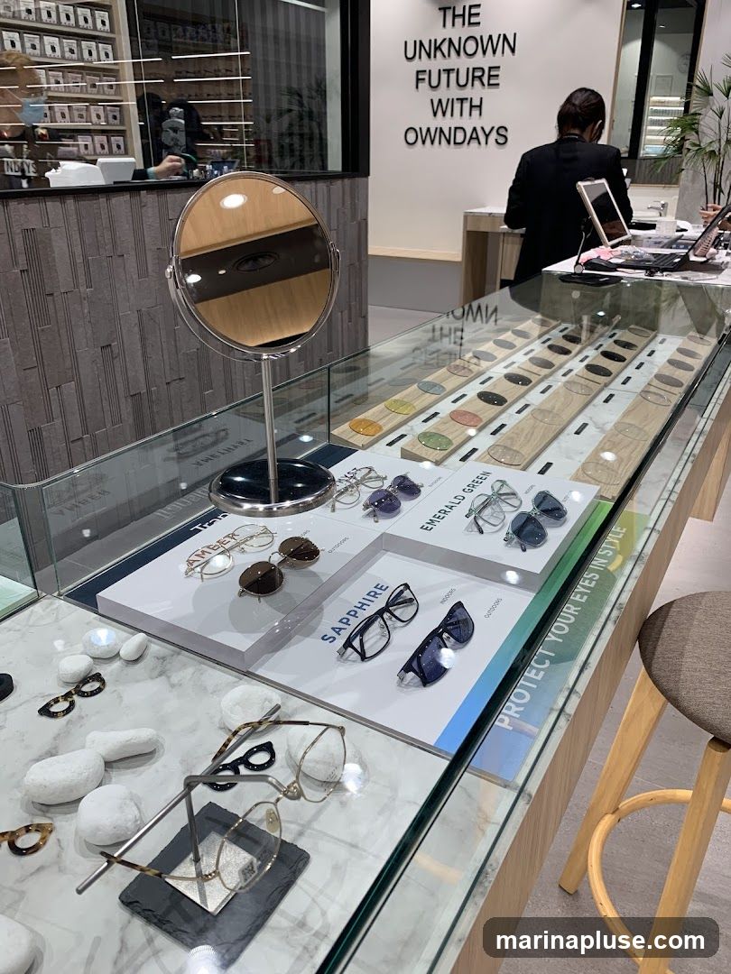 A display case filled with various eyeglasses and sunglasses. There are also samples of lenses in different colors. In the background, a sign reads "THE UNKNOWN FUTURE WITH OWNDAYS". A person in a black suit is visible behind a counter.