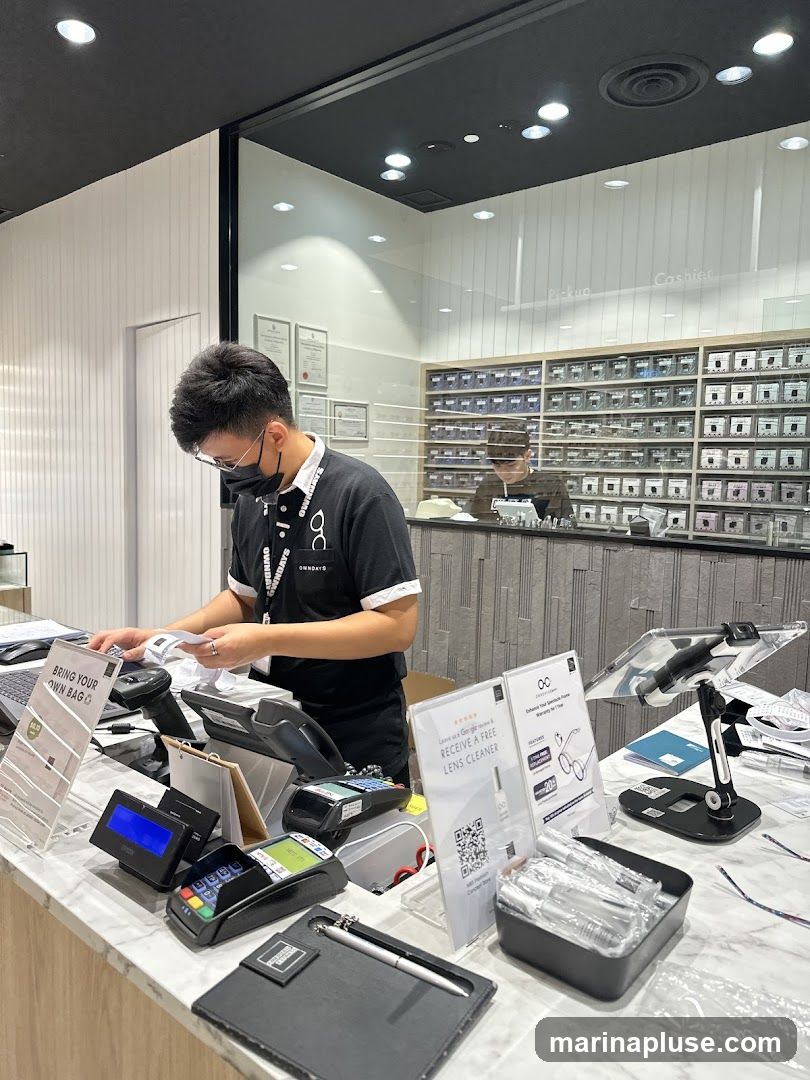 A staff member wearing a black shirt with a white collar and a lanyard is standing behind a counter, processing a transaction. The counter is equipped with a scanner, credit card machines, and a tablet. Behind the counter, shelves are filled with small boxes, likely containing eyewear or accessories. A sign on the counter advertises a free lens cleaner with a Google review.