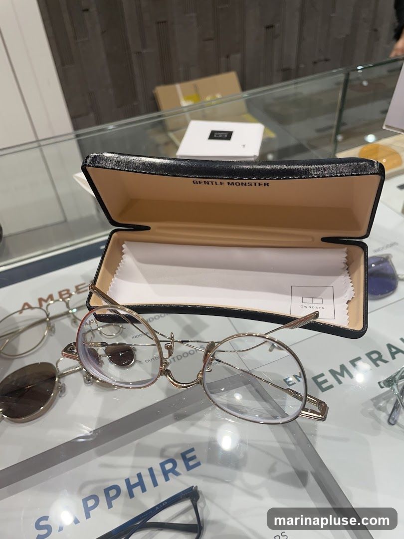 A pair of gold-rimmed eyeglasses rests on a glass surface, partially inside an open black glasses case. The inside of the case is lined with tan material and has the text "GENTLE MONSTER" printed on it. A white cleaning cloth with the text "OWNDAYS" is also visible inside the case. Other eyeglasses and labels such as "SAPPHIRE" and "EMERALD" are visible on the surface.