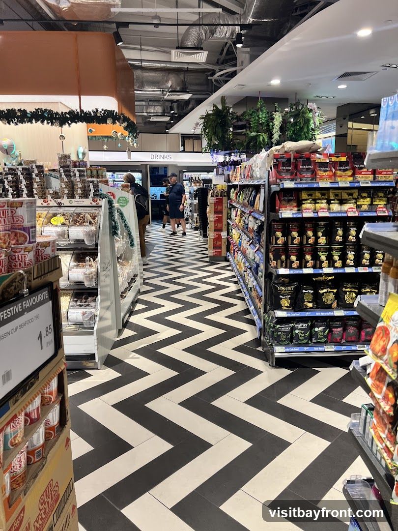 A view down an aisle in a supermarket with a distinctive black and white chevron patterned floor. Shelves stocked with various food items, including cup noodles, snacks, and drinks, line the sides of the aisle. Christmas decorations are visible in the background.