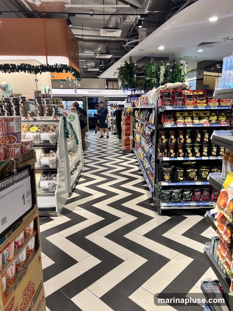 A view down an aisle in a supermarket with a distinctive black and white chevron patterned floor. Shelves stocked with various food items, including cup noodles, snacks, and drinks, line the sides of the aisle. Christmas decorations are visible in the background.
