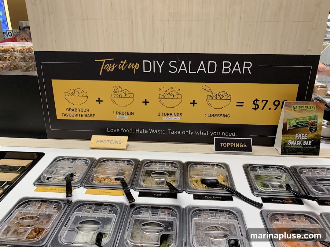 A salad bar with various ingredients in clear containers. A sign above explains the DIY salad bar concept: "Grab your favourite base, 1 protein, 2 toppings, 1 dressing" for $7.90. There is also a promotional sign for a free Nature Valley snack bar.