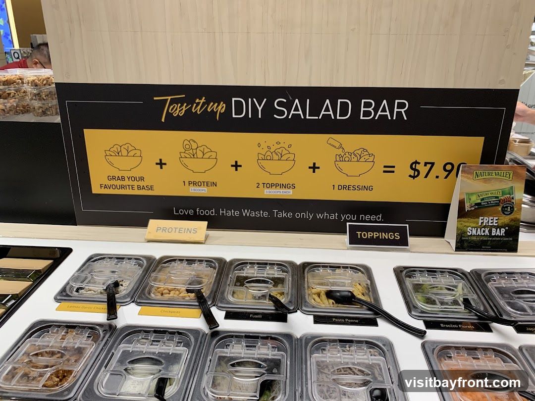 A salad bar with various ingredients in clear containers. A sign above explains the DIY salad bar concept: "Grab your favourite base, 1 protein, 2 toppings, 1 dressing" for $7.90. There is also a promotional sign for a free Nature Valley snack bar.