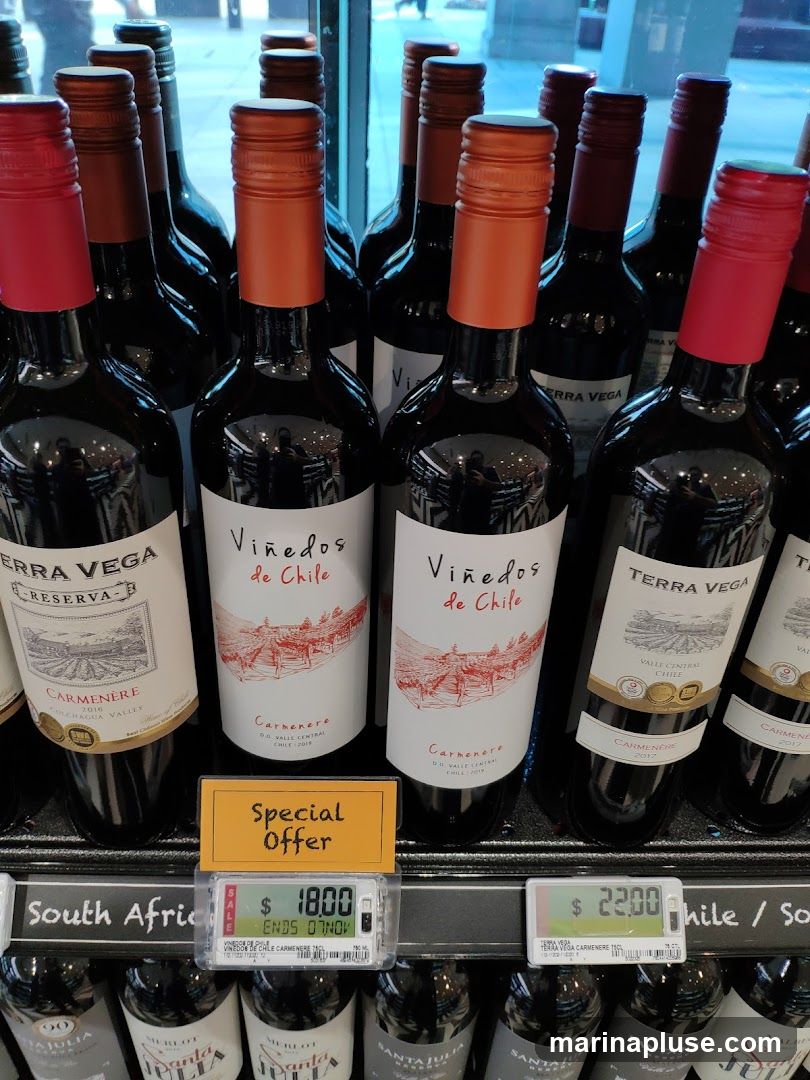 A selection of red wine bottles, primarily from Chile, are displayed on shelves. Some bottles have labels indicating "Viñedos de Chile" and "Terra Vega Reserva Carmenere". A "Special Offer" sign is visible, along with electronic price tags showing $18.00 and $22.00. The background suggests an indoor retail environment with reflections of people and store aisles.