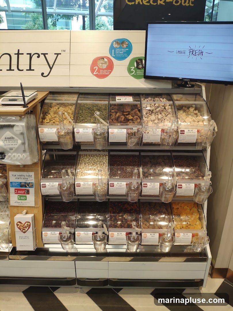 A display of bulk food bins filled with various dried fruits, nuts, and seeds. The bins are labeled with prices and product names. A sign above reads "ntry TM" and a screen displays "CHOOSE FRESH".