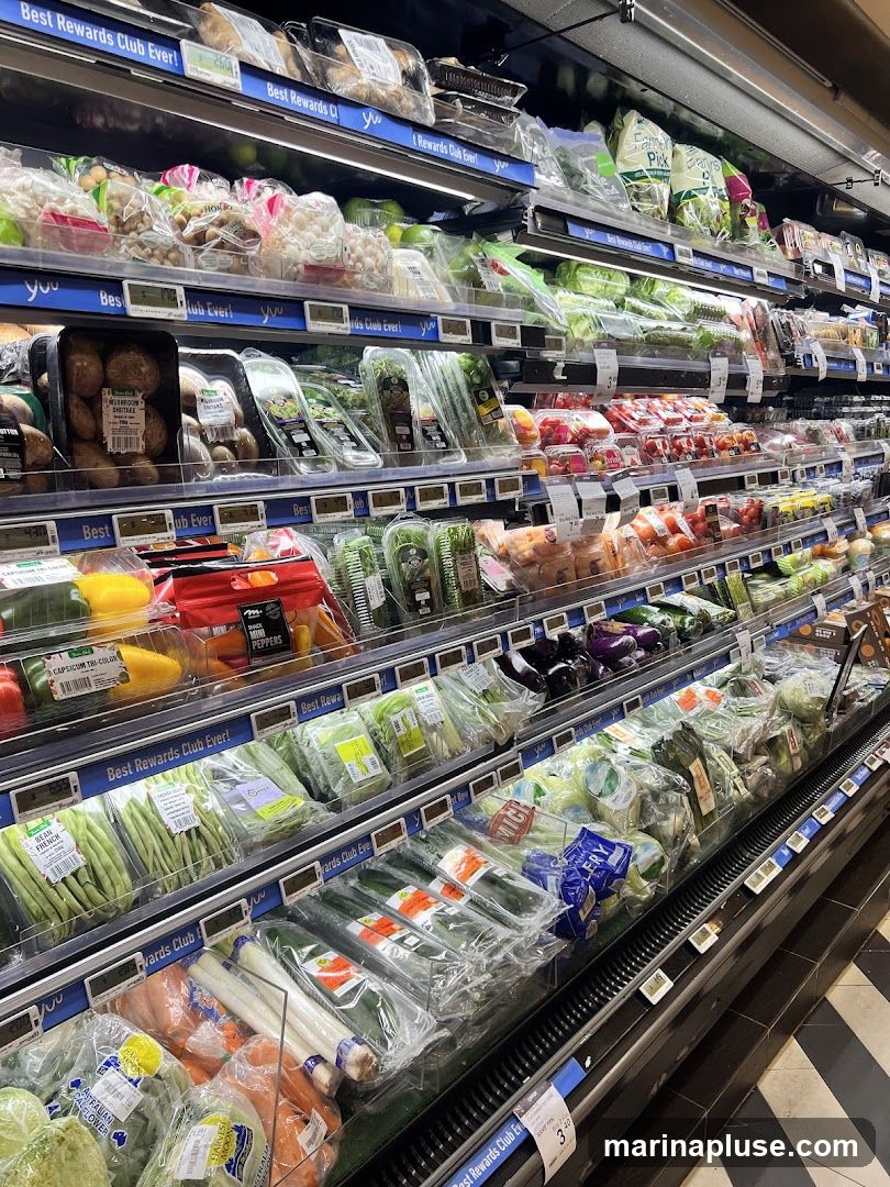 A refrigerated aisle in a grocery store displays a wide variety of fresh produce, including mushrooms, bell peppers, green beans, cucumbers, leeks, carrots, and various leafy greens. The items are neatly packaged and arranged on shelves, with price tags and "Best Rewards Club Ever!" signage visible.