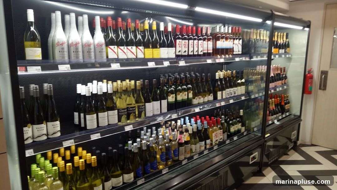 A refrigerated display case filled with a variety of wine bottles. The shelves are stocked with white, red, and rosé wines, as well as some sparkling wines and champagnes. Some bottles have visible labels, including Penfolds, Waiheke River, and Chanson. Prices are visible on some shelves. A red fire extinguisher is mounted on the wall to the right of the display. The floor has a black and white chevron pattern.