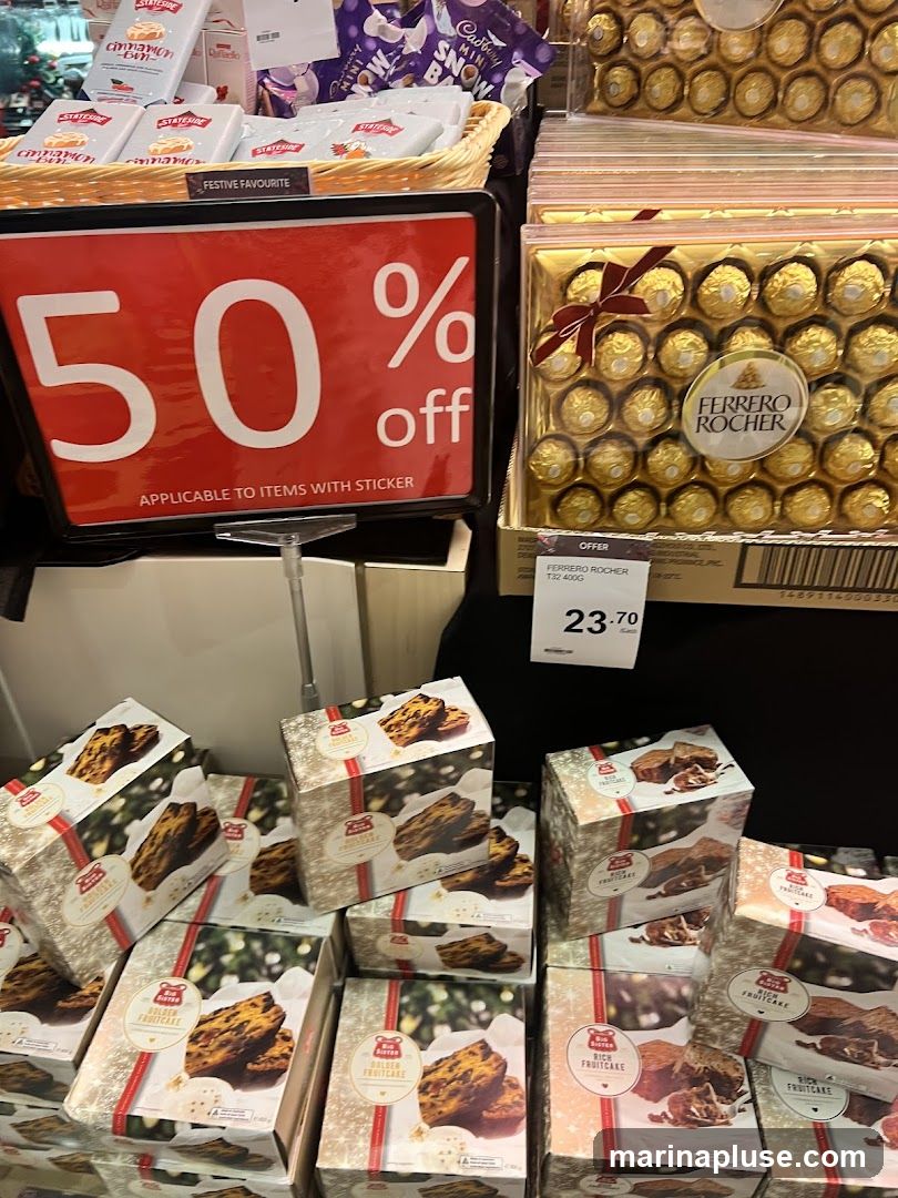 A display of Christmas-themed food items, including fruitcakes and Ferrero Rocher chocolates. A prominent sign advertises "50% off Applicable to Items with Sticker." Another sign shows Ferrero Rocher priced at 23.70.