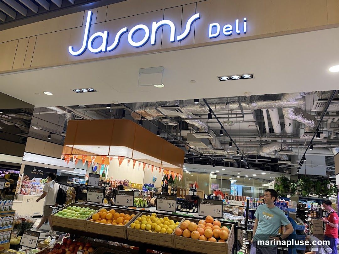 An interior view of Jason's Deli, a grocery store, with a prominent sign above the entrance. The store features displays of fresh fruits like apples, oranges, and lemons. Shelves stocked with various products, including drinks and packaged goods, are visible in the background. Several customers and staff members are present in the store.