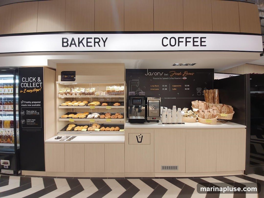 A counter with a display of baked goods and a coffee station. Above the counter, signs read "BAKERY" and "COFFEE". To the left, a display cooler contains drinks, and a sign advertises "CLICK & COLLECT". The coffee station features a machine, cups, and a menu board for "Jasons Deli Fresh Brews" with prices for Long Black, Americano, Cappuccino, and Latte. Baskets of bread are also visible.