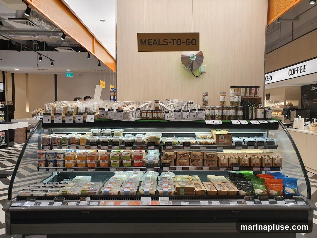 A refrigerated display case filled with various pre-packaged meals and food items. Above the case, a sign reads "MEALS-TO-GO." In the background, there are signs for "BAKERY" and "COFFEE." The items in the case include salads, sandwiches, and other ready-to-eat options, with some prices visible.