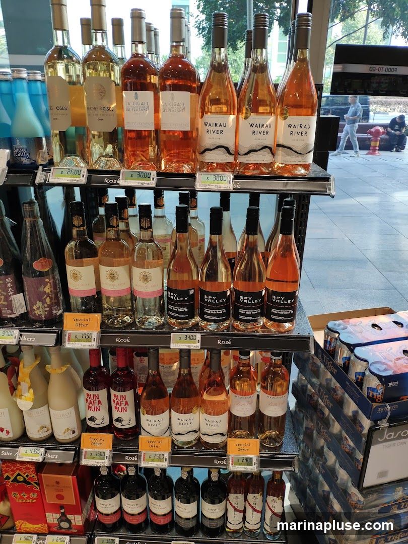 A retail display featuring several shelves of wine bottles, primarily rosé, along with some other beverages. Price tags are visible for many of the bottles. In the background, a street scene with people and a fire hydrant can be seen through a large window.