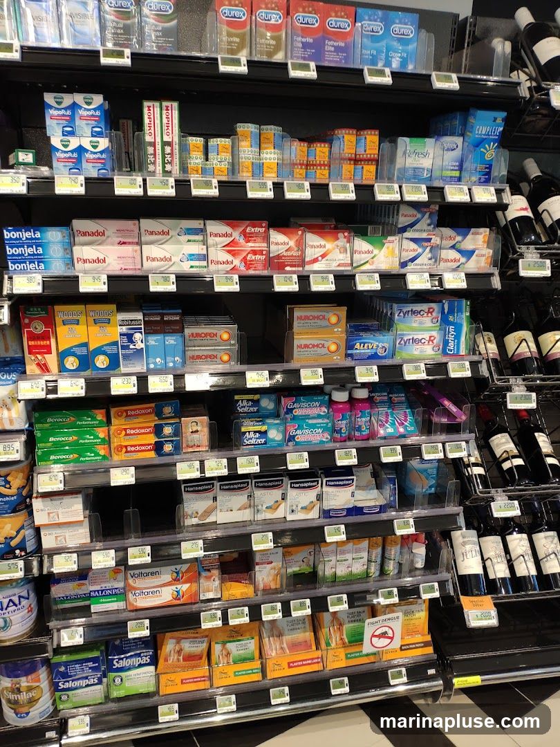 A retail display of various health and personal care products on multiple shelves. Products include medications like Panadol, Nurofen, and Voltaren, as well as personal hygiene items such as Durex condoms and VapoRub. There are also vitamins, pain relief patches, and some bottled beverages, possibly wine, on the right side. Digital price tags are visible below many items.