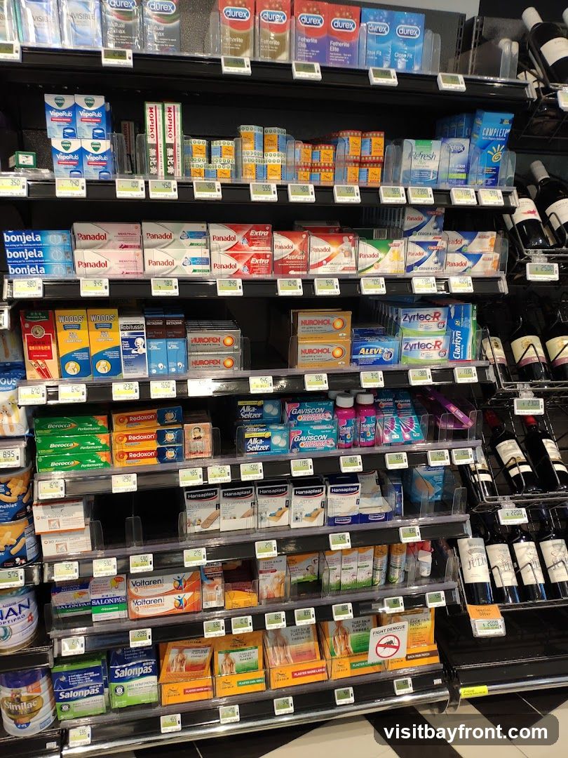 A retail display of various health and personal care products on multiple shelves. Products include medications like Panadol, Nurofen, and Voltaren, as well as personal hygiene items such as Durex condoms and VapoRub. There are also vitamins, pain relief patches, and some bottled beverages, possibly wine, on the right side. Digital price tags are visible below many items.